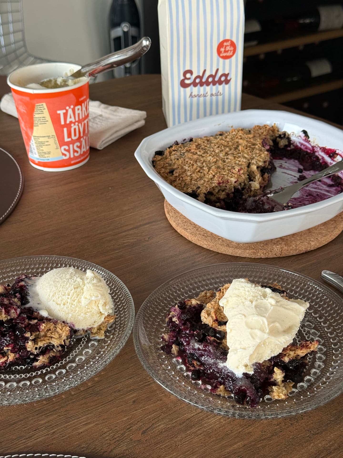 Bilberry oat crumble served on glass plates with vanilla ice cream, next to a bag of Edda honest oats
